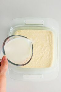 A hand pours milk from a glass measuring cup into a square glass dish filled with batter.