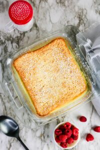 A baked square cake dusted with powdered sugar in a glass dish on a marble surface, next to a bowl of raspberries, a spoon, and a sugar shaker.