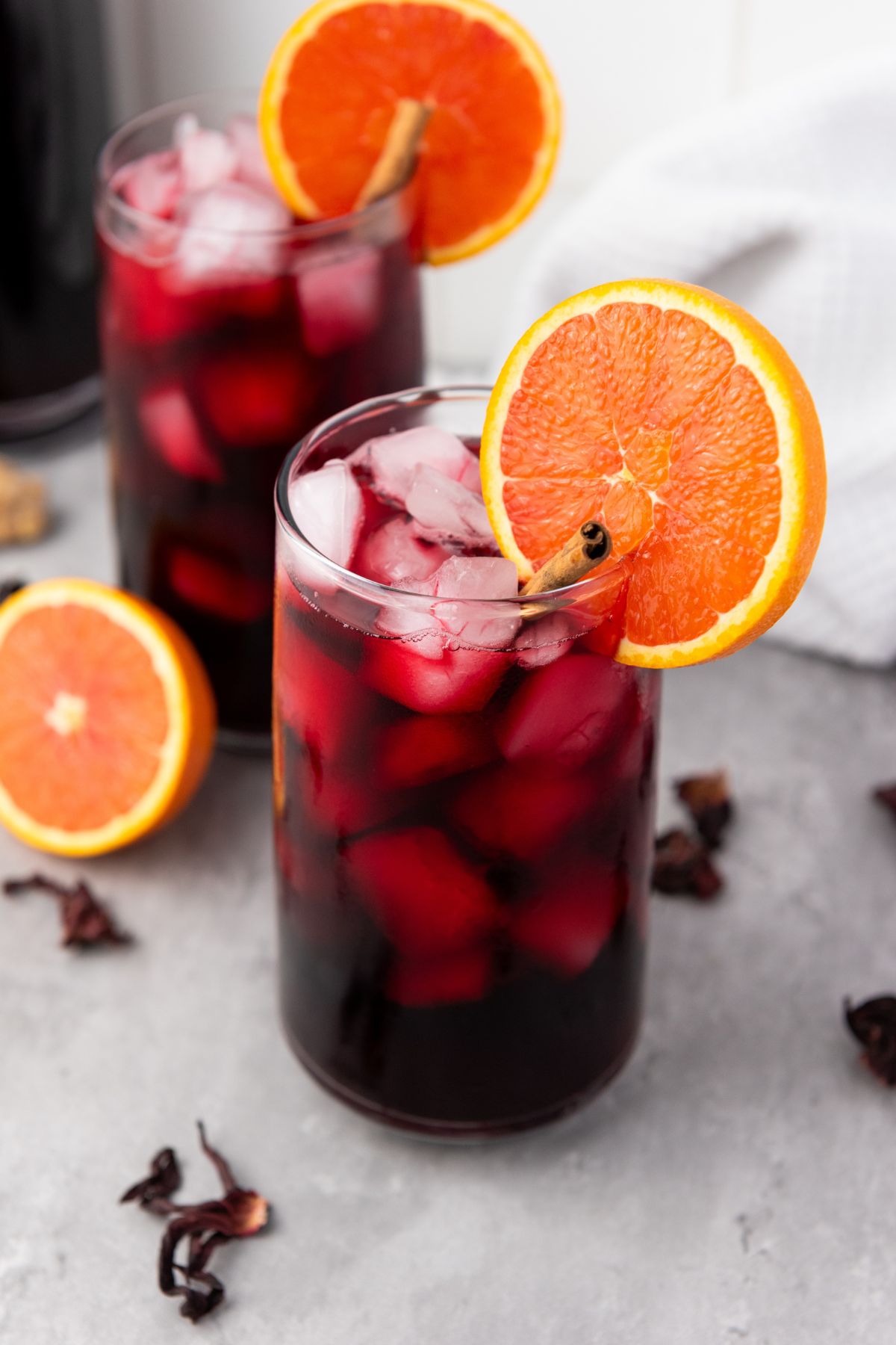 Two glasses of iced hibiscus drink garnished with orange slices, placed on a gray surface with scattered dried hibiscus petals and orange slices nearby.