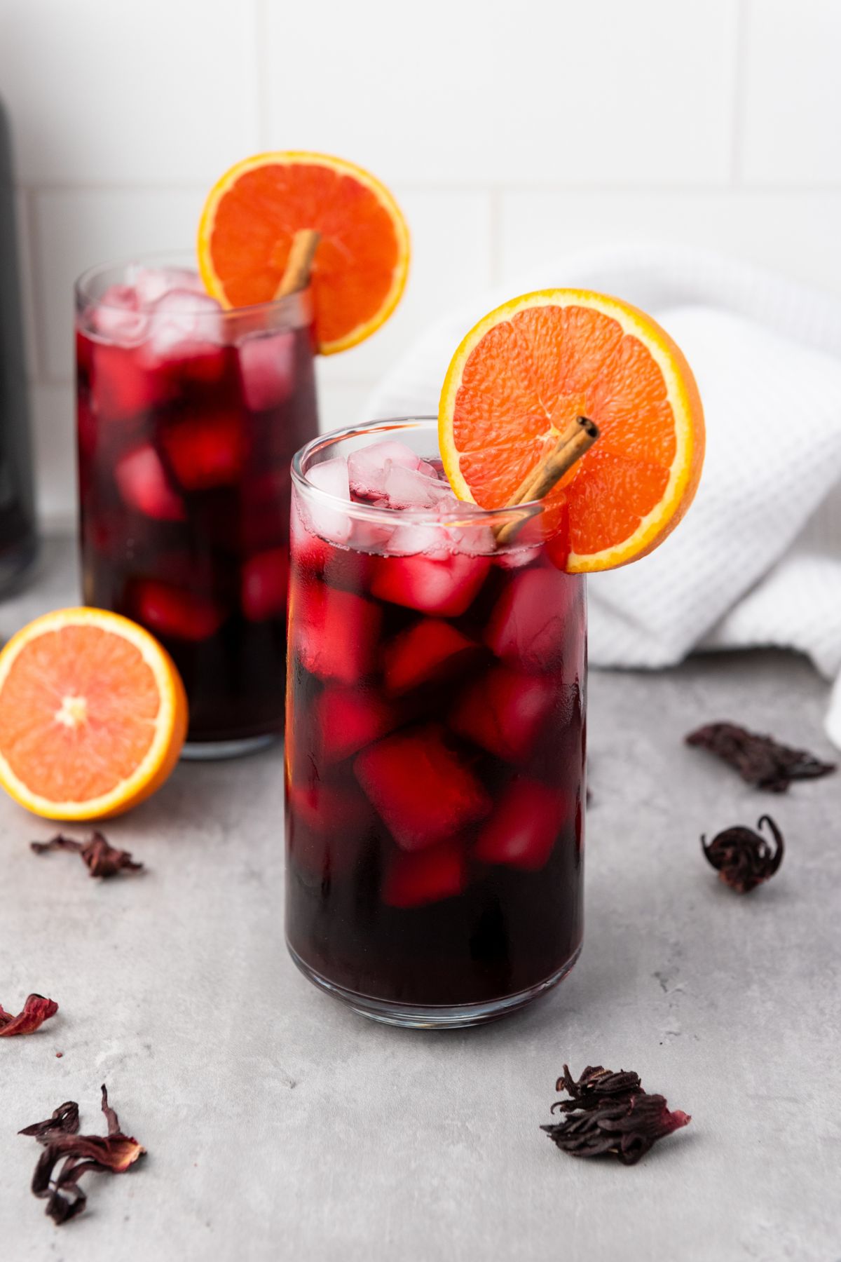 Two glasses of iced red beverage garnished with orange slices and cinnamon sticks, with dried hibiscus flowers and an orange half on a gray surface.