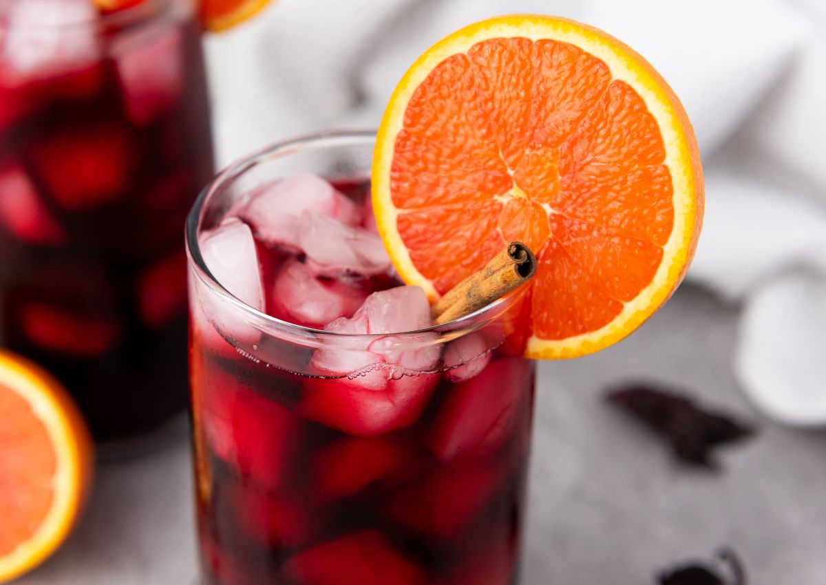 A glass of iced red drink garnished with a slice of orange and a cinnamon stick, with another similar glass blurred in the background.