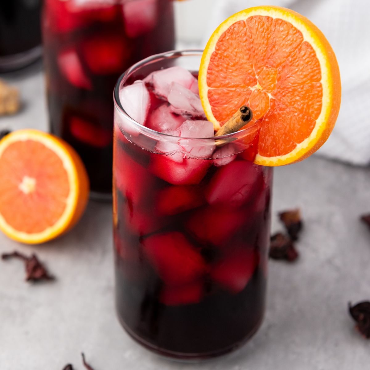 A glass of iced red hibiscus drink garnished with an orange slice and a cinnamon stick, with another glass and orange slices in the background.