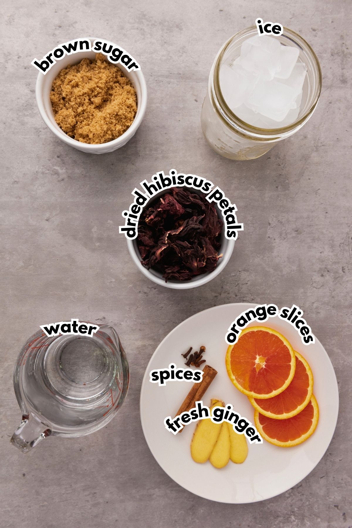 Bowls and plates with brown sugar, dried hibiscus petals, ice, water, spices, orange slices, and fresh ginger are arranged on a gray surface, each labeled with text.