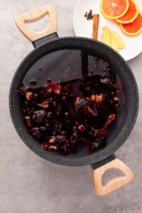 A black pot with wooden handles contains dark liquid with hibiscus flowers. Beside the pot, a plate holds orange slices, ginger, and a cinnamon stick.