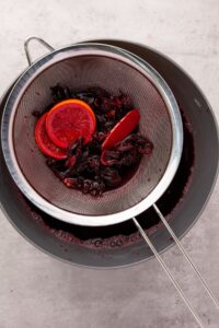 A metal strainer with hibiscus petals and orange slices rests over a bowl, filtering dark red liquid.