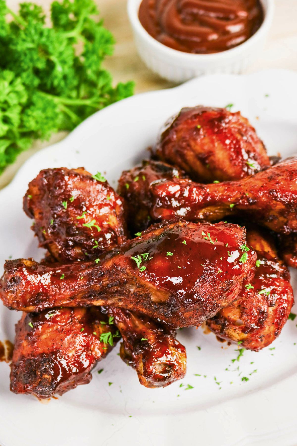 Barbecue-glazed chicken drumsticks arranged on a white plate, garnished with chopped herbs, with a cup of barbecue sauce and parsley in the background.