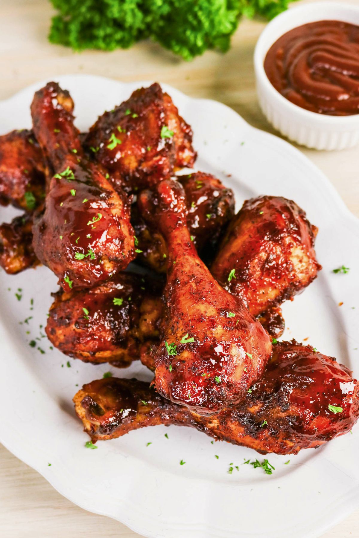 Barbecued chicken drumsticks coated in sauce are arranged on a white plate, garnished with chopped herbs. A small bowl of barbecue sauce and leafy greens appear in the background.
