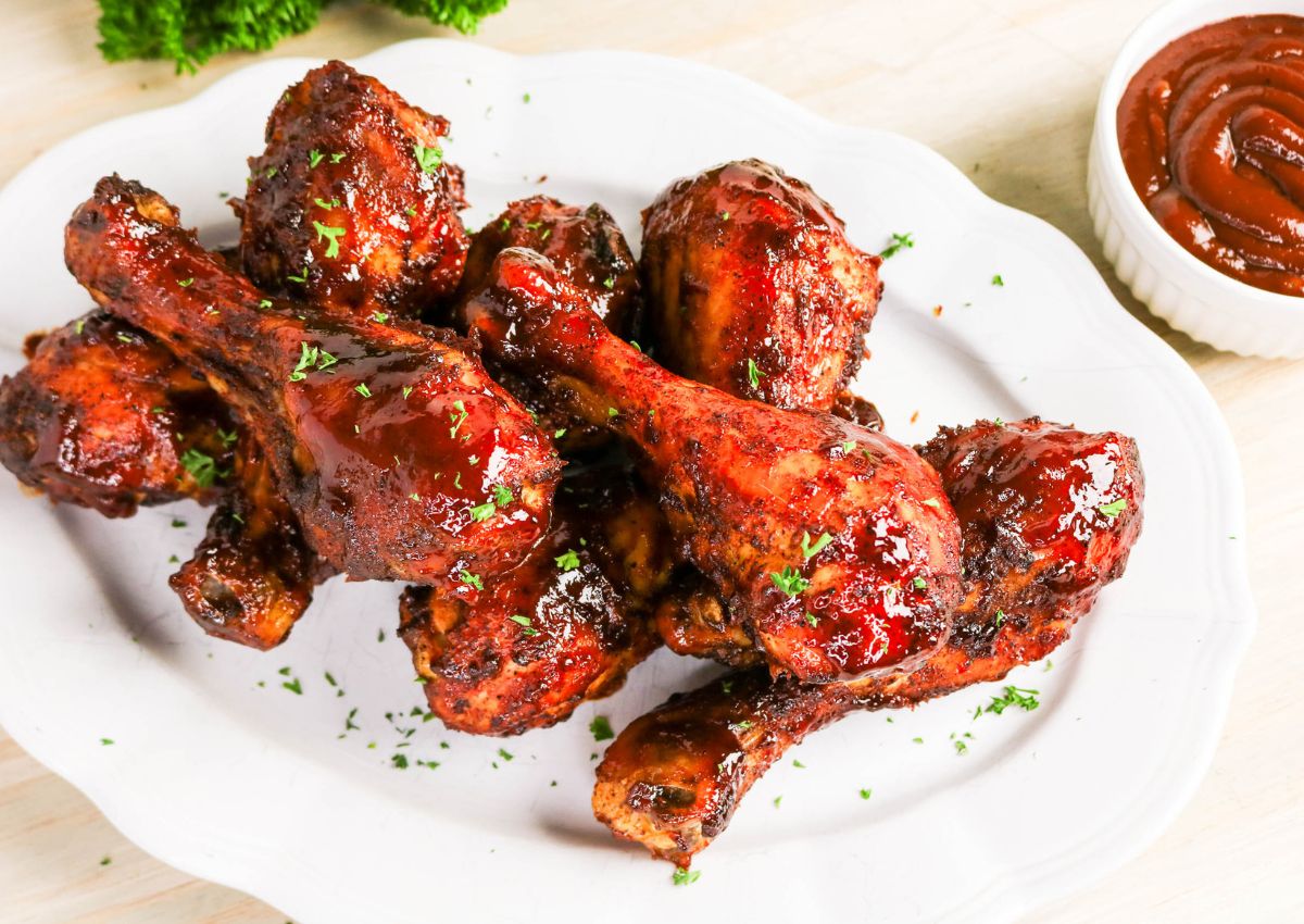 A white plate with several glazed barbecue chicken drumsticks, sprinkled with chopped herbs, next to a small bowl of barbecue sauce.