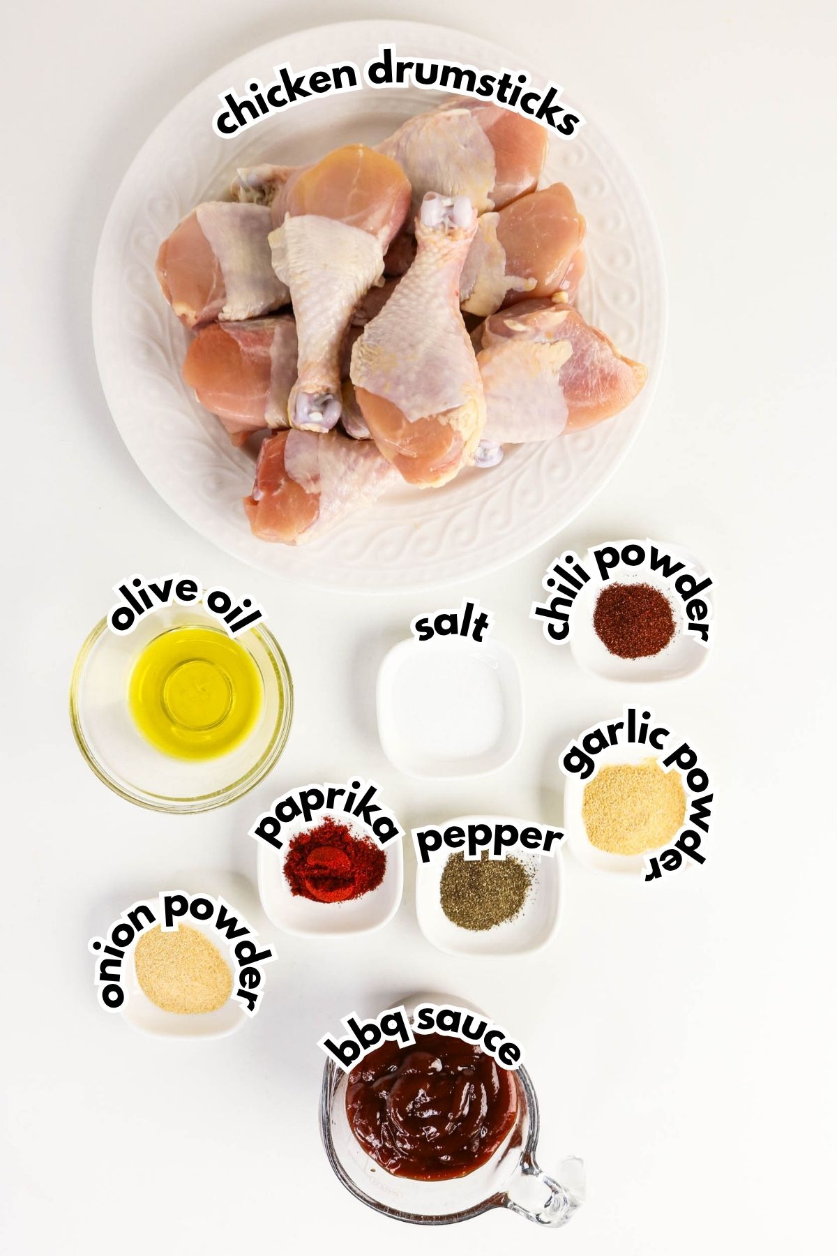 A plate of raw chicken drumsticks with small bowls of olive oil, salt, chili powder, garlic powder, pepper, onion powder, paprika, and bbq sauce arranged on a white surface.
