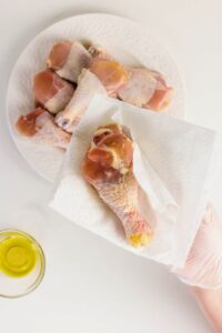 A hand in a glove uses a paper towel to pat dry a raw chicken drumstick over a plate of chicken pieces. A small bowl of oil is nearby.