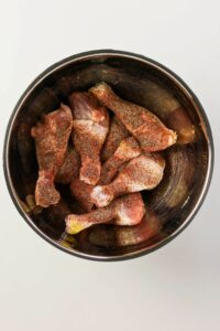 Raw chicken drumsticks coated with spices in a metal mixing bowl, placed on a plain white surface.