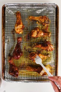 Chicken drumsticks on a wire rack over a foil-lined baking sheet, with a hand brushing sauce onto one drumstick using a silicone brush.