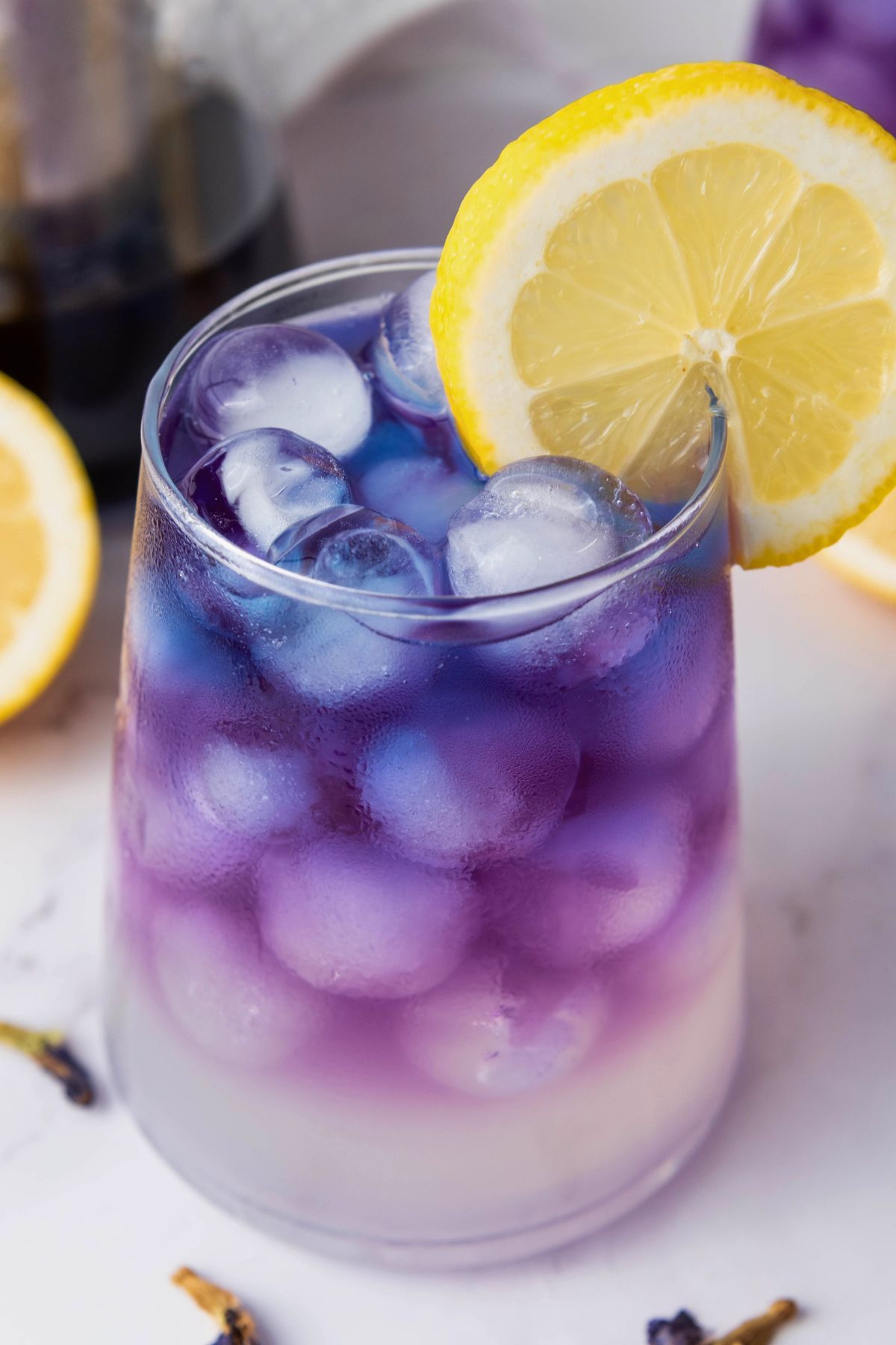 A glass filled with ice cubes and a layered purple and white beverage, garnished with a lemon slice on the rim.