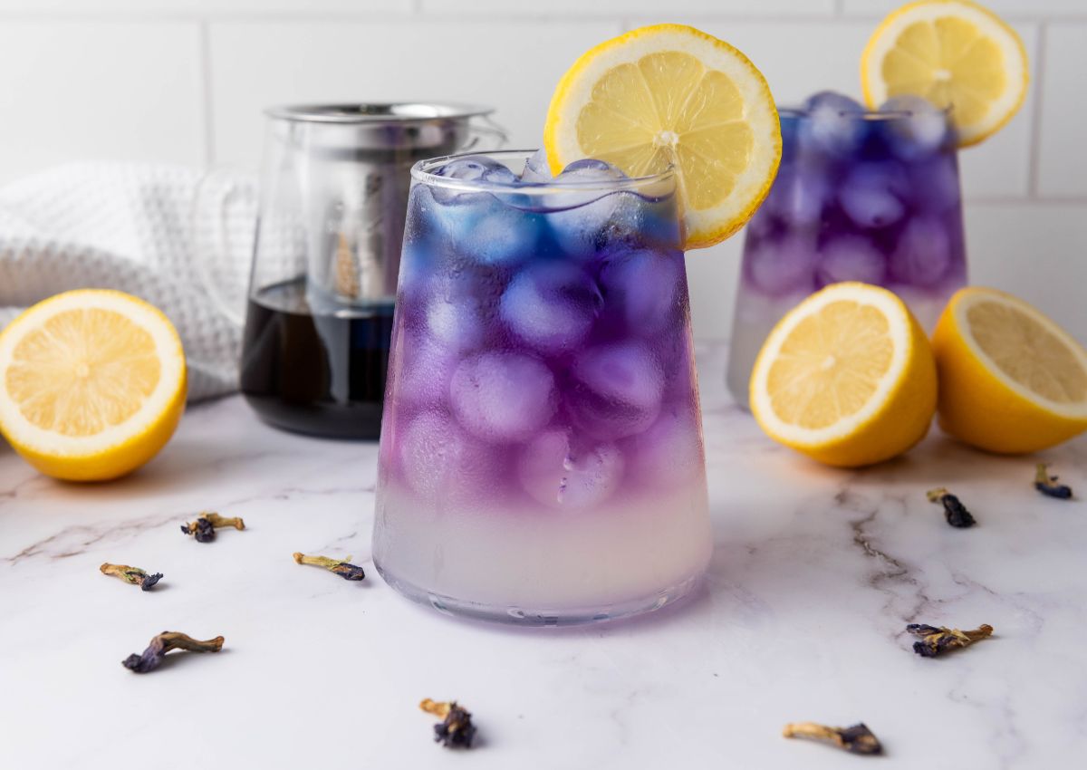 A glass of iced butterfly pea lemonade with a gradient from purple to white, garnished with a lemon slice; whole and sliced lemons and butterfly pea flowers surround the glass.