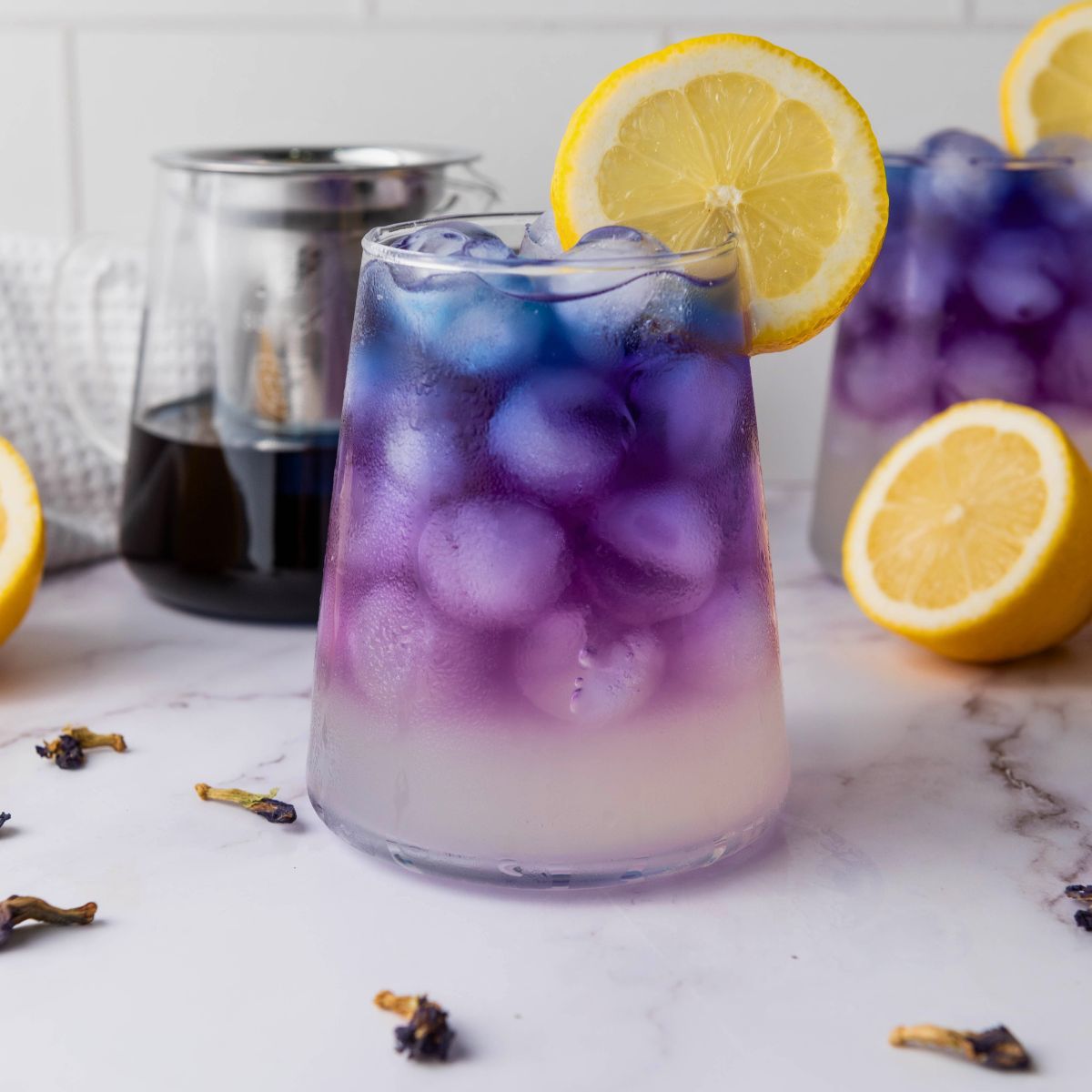 A glass of iced purple lemonade with a lemon slice garnish sits on a marble surface, surrounded by lemon halves and a glass pitcher with dark liquid.