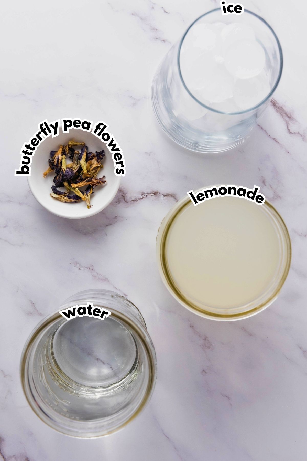 A glass of ice, a bowl of dried butterfly pea flowers, a jar of water, and a glass of lemonade are arranged on a marble surface with text labels for each item.