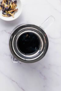 A glass teapot filled with dark blue liquid sits on a marble surface beside a small bowl of dried butterfly pea flowers.
