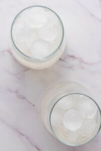 Two round glasses filled with clear liquid and ice cubes on a light marble surface, viewed from above.