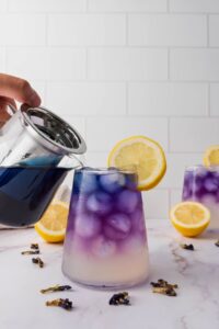 A hand pours blue liquid from a teapot into a glass of iced lemonade, causing the drink to change color; lemon slices and scattered petals are on the table.