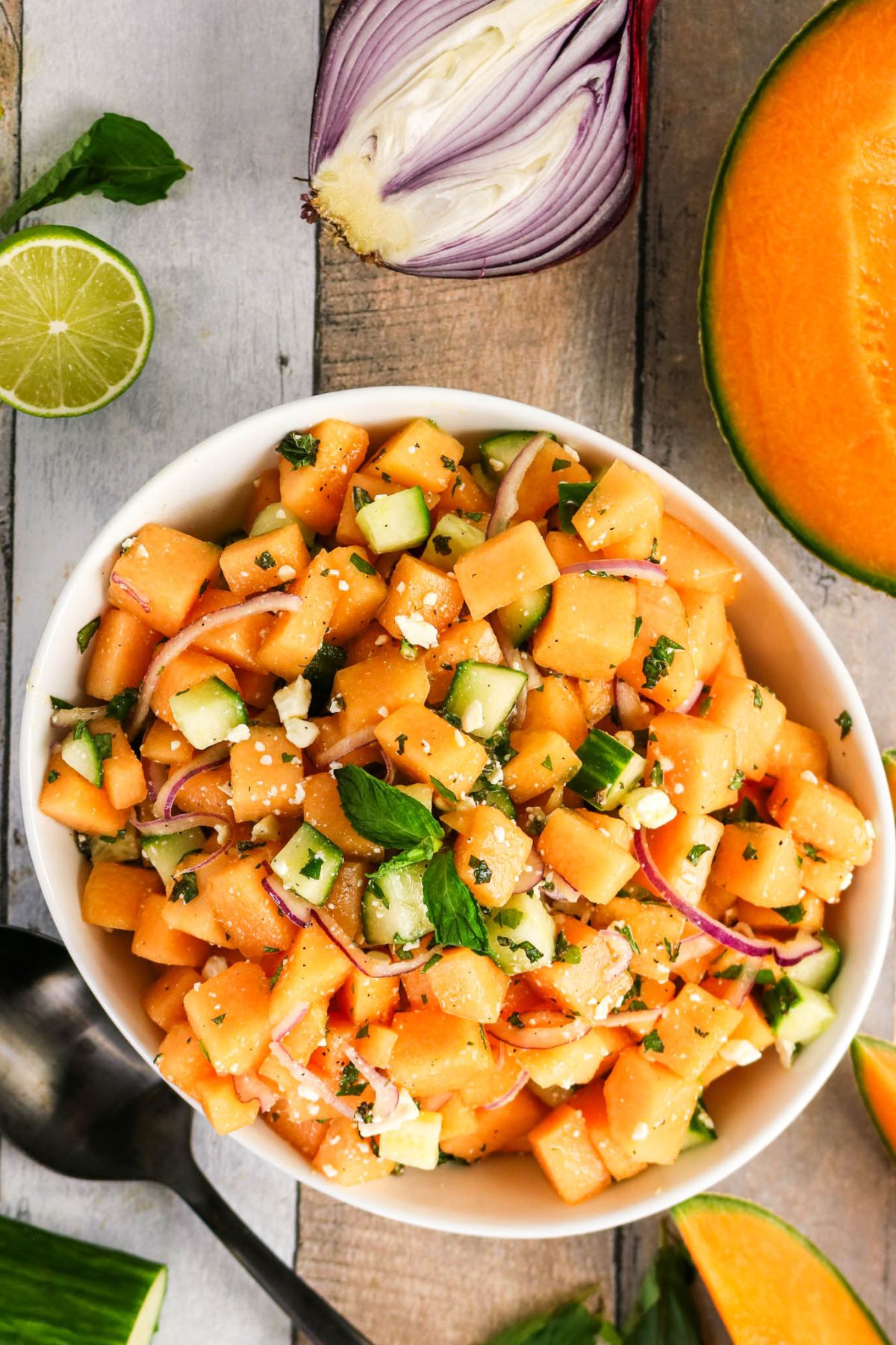 A bowl of cantaloupe salad with cucumber, red onion, feta cheese, and fresh herbs, surrounded by ingredients including cantaloupe, lime, and red onion on a wooden surface.