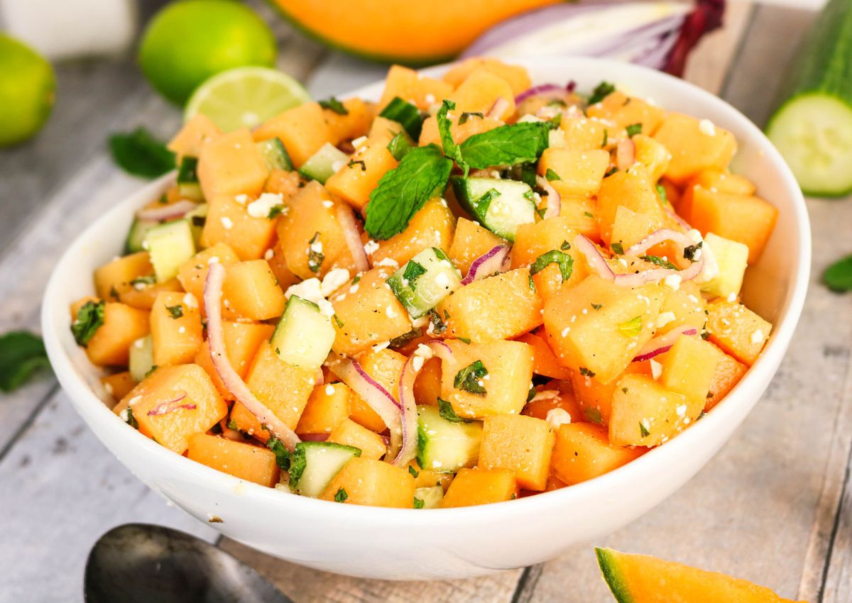 A bowl of cantaloupe and cucumber salad with red onion, mint, and feta cheese on a wooden surface, garnished with lime and mint leaves.