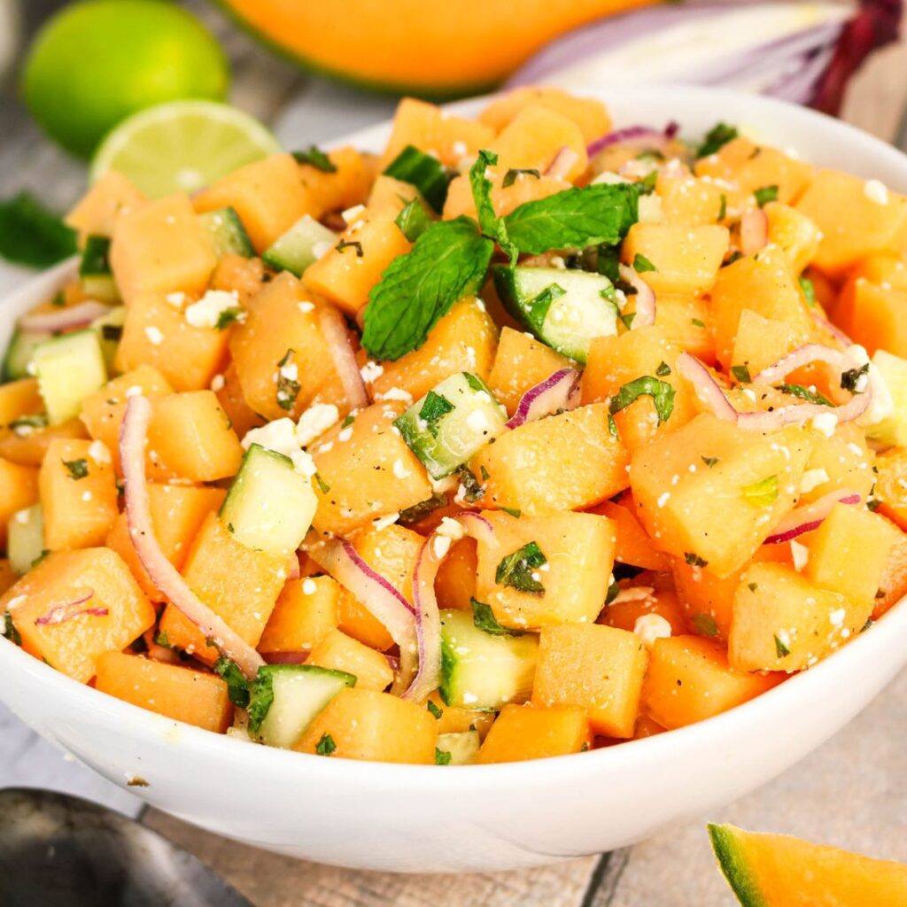 A bowl of cantaloupe salad with diced cantaloupe, cucumber, red onion, feta cheese, and fresh mint leaves, garnished with a sprig of mint.