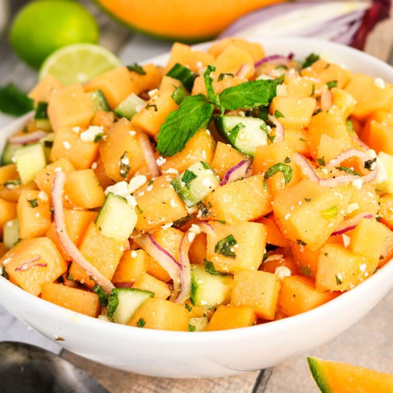A bowl of cantaloupe salad with diced cantaloupe, cucumber, red onion, feta cheese, and fresh mint leaves, garnished with a sprig of mint.