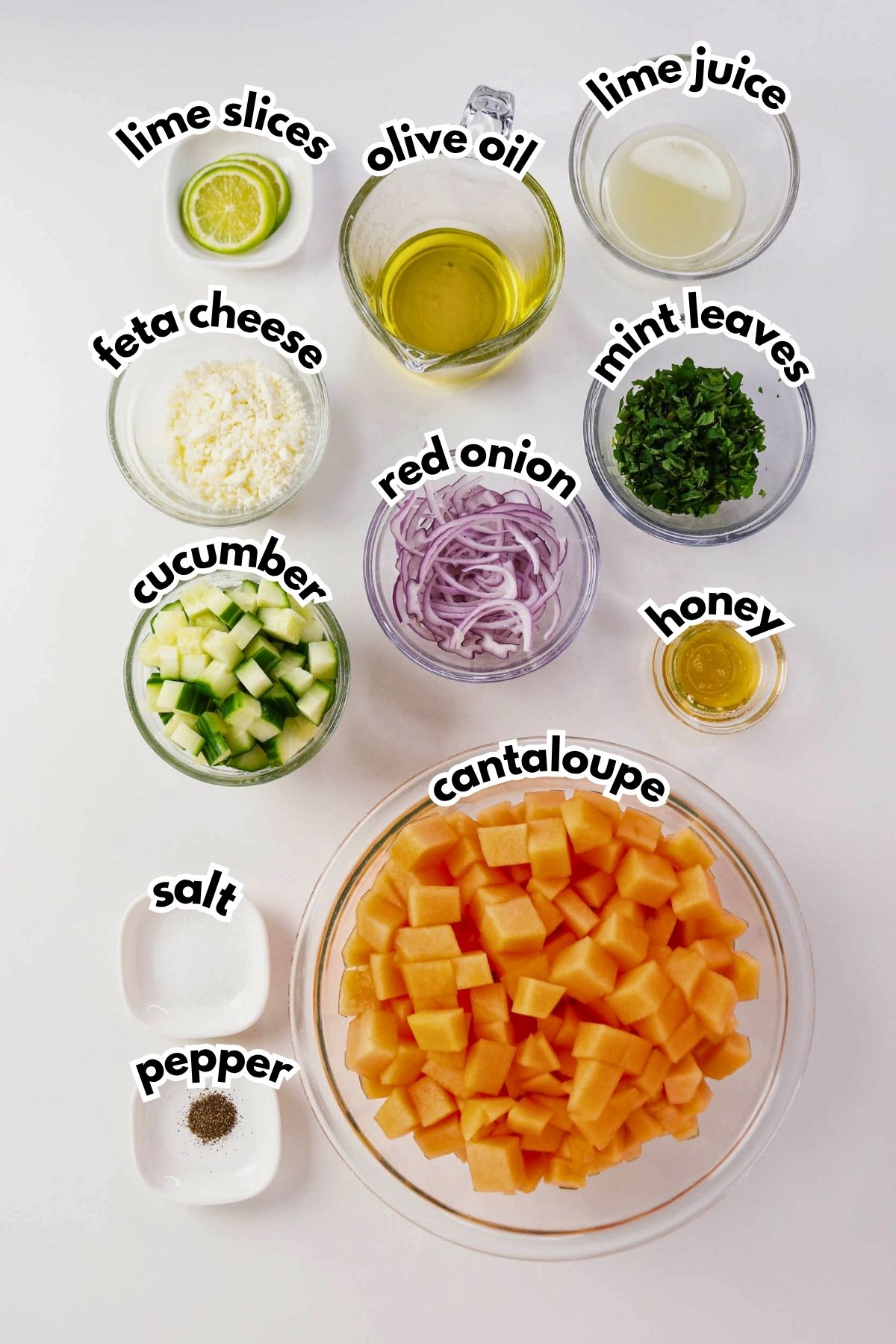 Bowls of diced cantaloupe, cucumber, red onion, feta cheese, chopped mint, lime slices, olive oil, lime juice, honey, salt, and pepper arranged on a white surface.