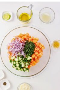 A glass bowl with diced papaya, cucumbers, red onion, and chopped herbs on a white surface, surrounded by small bowls of olive oil, lemon juice, seasonings, and lime slices.