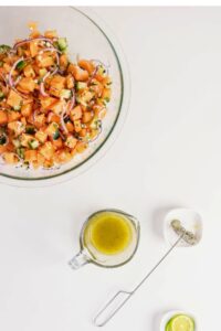 A bowl of chopped cantaloupe salad with red onion and cucumber, next to a small pitcher of dressing, a whisk, and a lime wedge on a white surface.