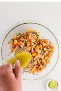 A hand pours yellow vinaigrette dressing over a bowl of chopped cantaloupe, cucumber, red onion, and herbs; a lime wedge is nearby.