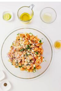 A glass bowl with chopped cantaloupe, cucumber, red onion, feta, and herbs, surrounded by small bowls of oil, lemon juice, pepper, and a halved lime on a white surface.