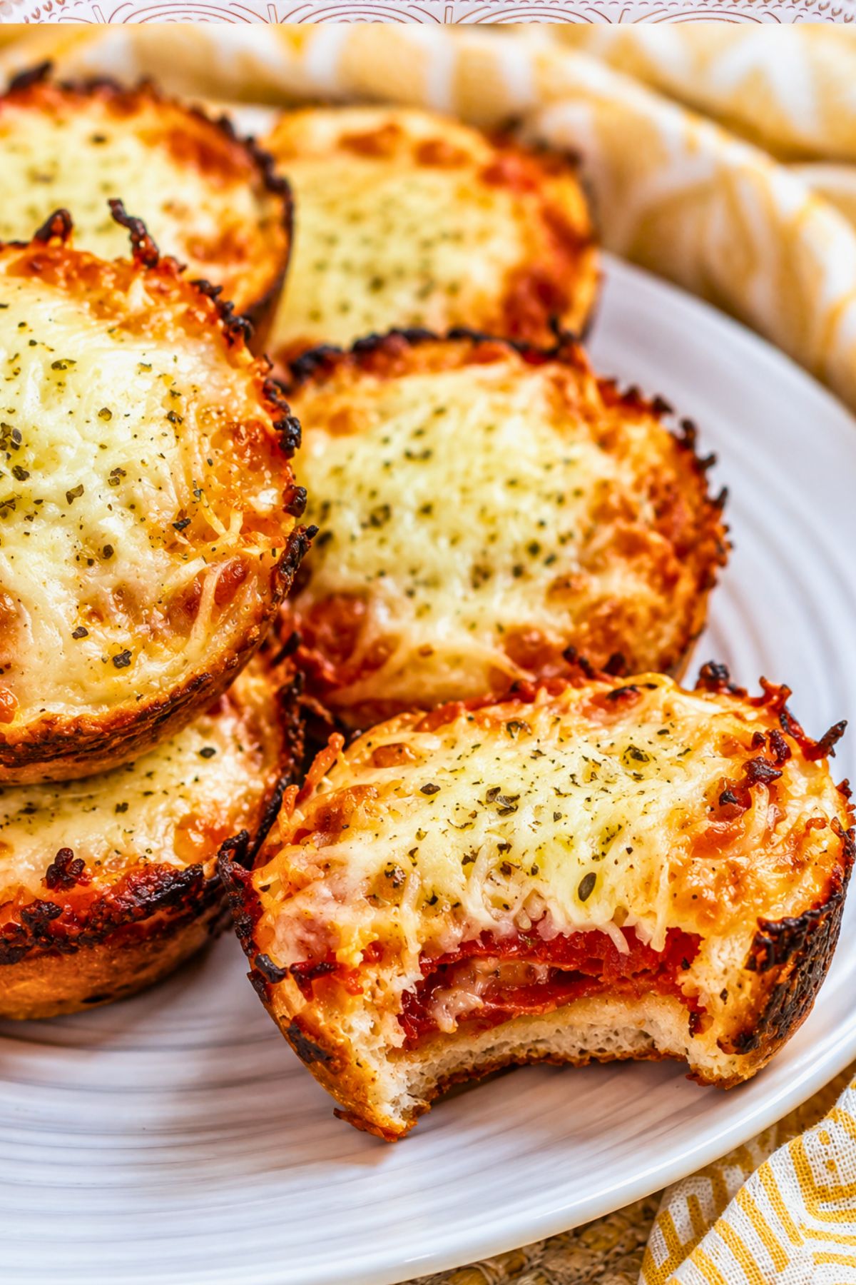A plate of mini pizza muffins topped with melted cheese and herbs, with one muffin showing a bite taken out to reveal layers of sauce and pepperoni.