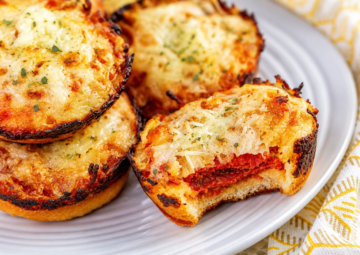 Several mini pizzas with melted cheese and herbs sit on a white plate; one pizza shows a visible bite, revealing layers of sauce and pepperoni.