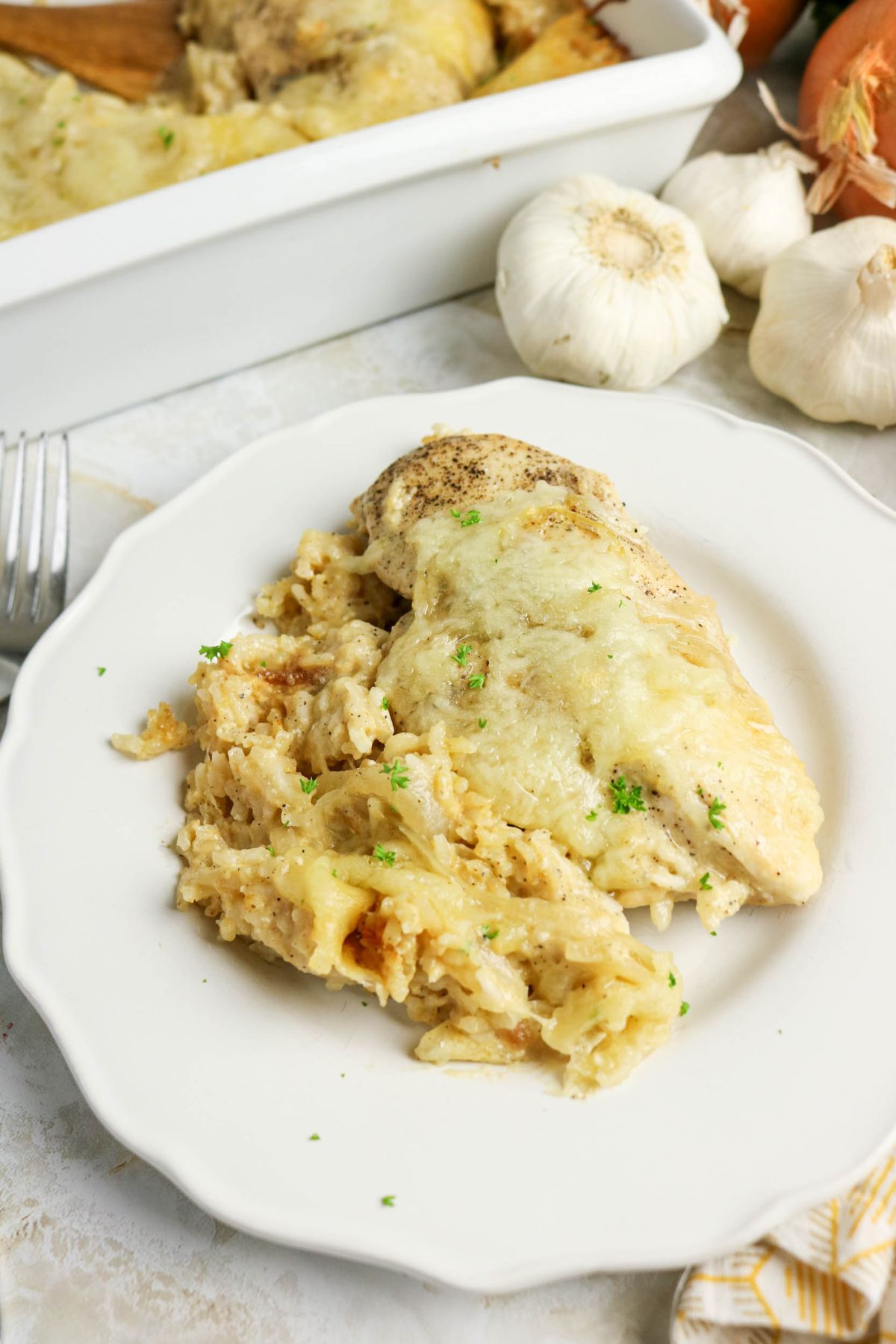Baked chicken breast with melted cheese served alongside cheesy rice on a white plate, with garlic bulbs and a casserole dish in the background.
