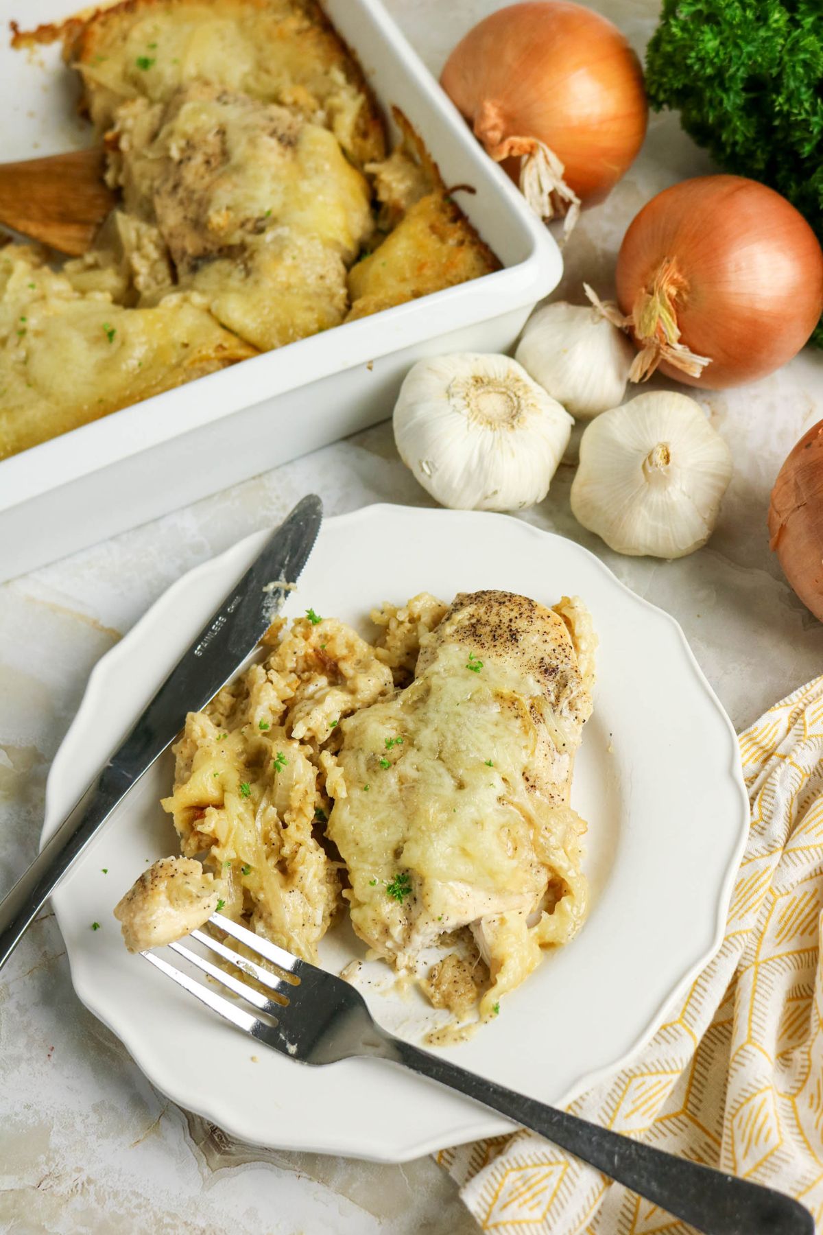 A serving of cheesy chicken casserole on a white plate with a fork and knife, next to onions, garlic, parsley, and a baking dish of casserole.