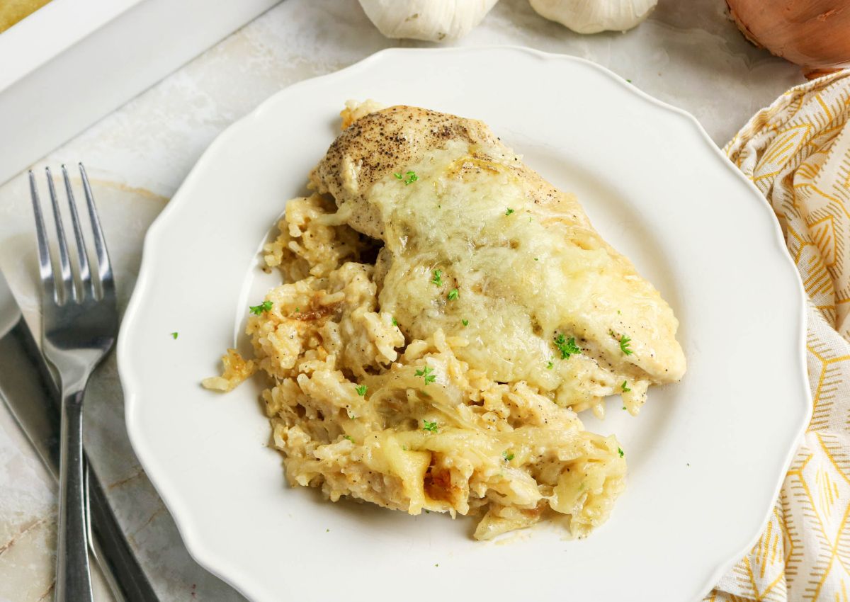 A cooked chicken breast topped with melted cheese, served over a creamy rice mixture on a white plate, with a fork and knife nearby.