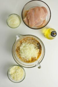 Five bowls and containers hold raw chicken breasts, sliced onions, olive oil, shredded cheese, and a mixing bowl with cheese, seasonings, and liquid ingredients on a white surface.