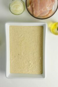 Rectangular dish filled with a creamy, seasoned batter. Surrounding it are bowls of raw chicken, sliced onions, and a small bottle of oil on a white surface.