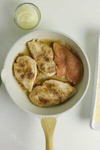 Four seasoned chicken breasts cooking in a white skillet, with a small bowl of sauce above and a white dish partially visible on the right.