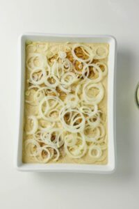 White baking dish filled with sliced onions layered over a creamy sauce and seasoned mixture, photographed from above on a white background.