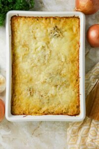 A baked casserole with a golden, melted cheese topping in a white rectangular dish, surrounded by onions, garlic, parsley, and a patterned cloth.