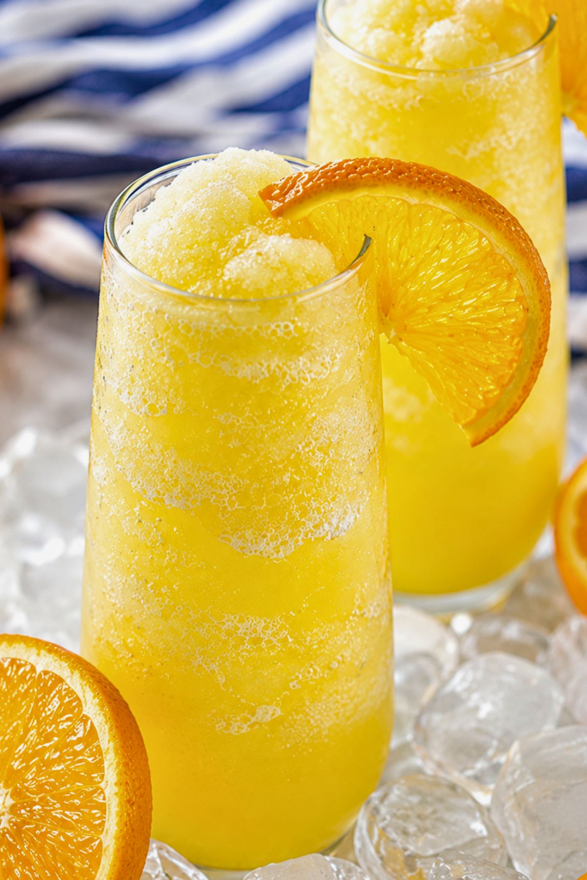 A tall glass of yellow slushy drink garnished with an orange slice, placed on ice cubes with a blue and white striped cloth in the background.