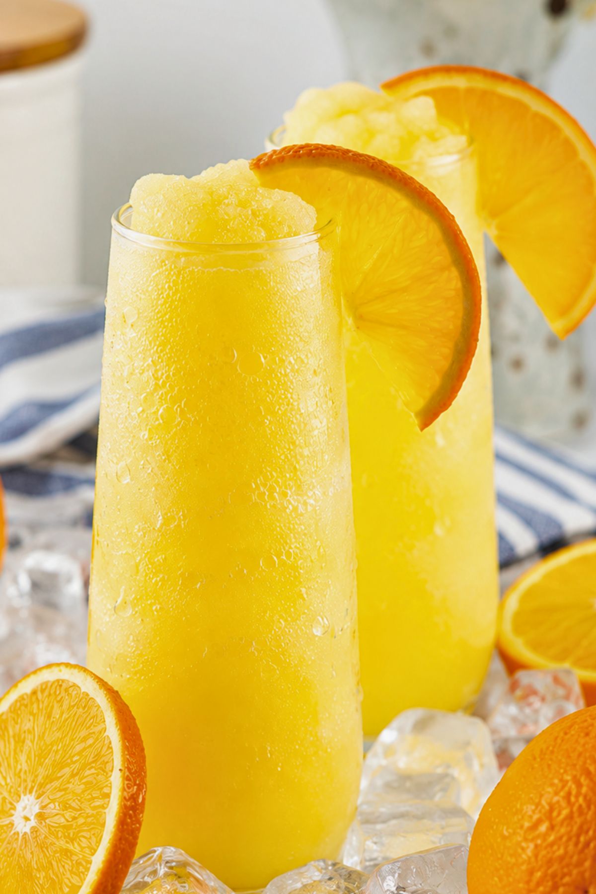 Two tall glasses of frozen orange slush garnished with orange slices, surrounded by ice cubes and fresh orange halves.