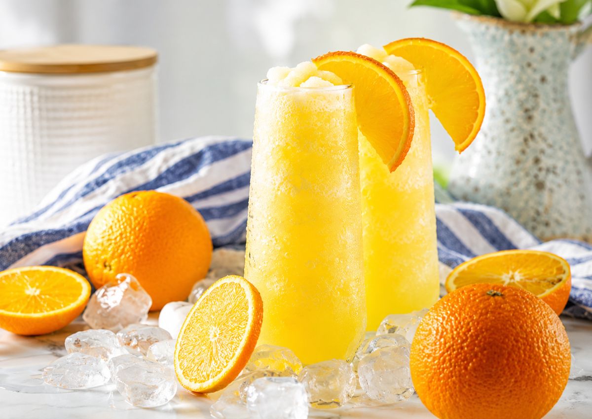 Two tall glasses of orange slush topped with orange slices, surrounded by ice cubes and whole and halved oranges on a white surface.
