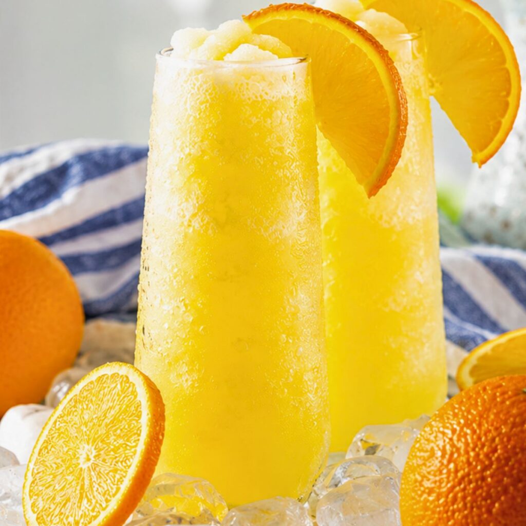 Two tall glasses of frozen orange drink garnished with orange slices, surrounded by ice cubes and whole oranges.