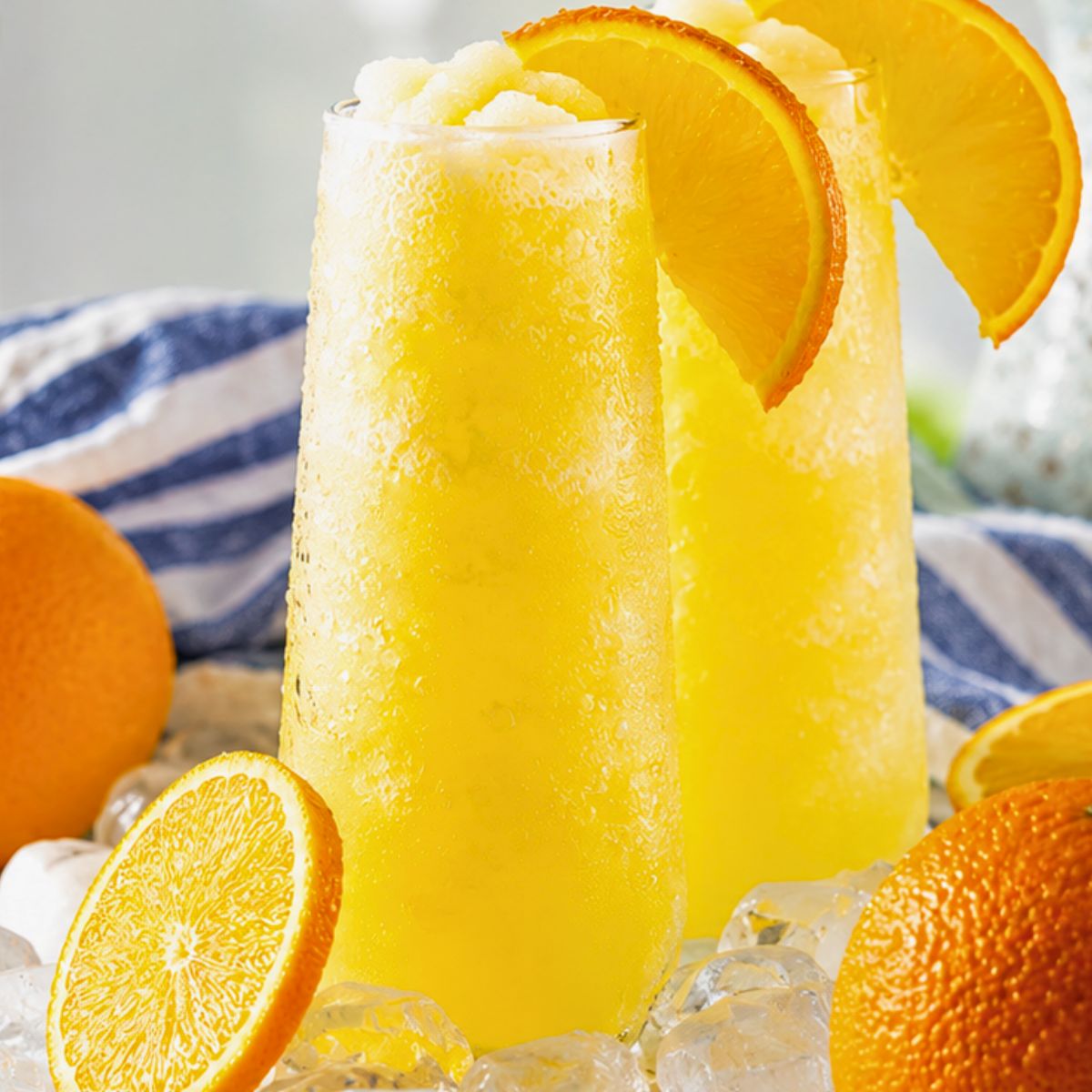 Two tall glasses of frozen orange drink garnished with orange slices, surrounded by ice cubes and whole oranges.