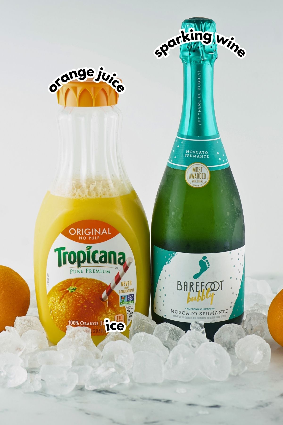 A bottle of Tropicana orange juice and a bottle of Barefoot Bubbly Moscato Spumante sparkling wine on ice, with oranges in the background.