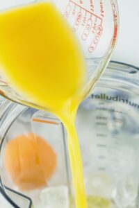 A measuring cup pours orange juice into a blender containing ice cubes and an egg.
