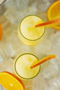 Two glasses of yellow frozen drink, each garnished with a slice of orange, are placed on a surface covered with ice and orange slices.